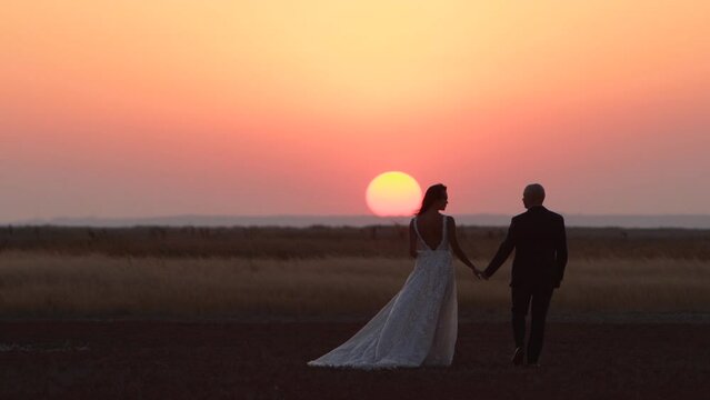 Newlywed couple holding hands, going to the big red sun at summer sunset. Just married man, woman walking across the nature outside at sunrise. Happy wife and husband posing at wedding photoshoot.