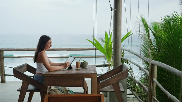 Young illustrator drawing on tablet at cafe with ocean view, palm trees. Pretty focused woman works remotely at authentic bar near tropical coast. Freelancer editing illustration at the seaside.