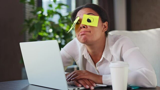Funny lazy multicultural woman blowing with plump lips on yellow sticky notes covering her eyes at workplace. Unmotivated company member in formal wear feeling tired from monotonous office work.