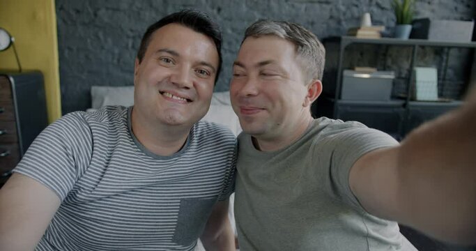 Portrait of adorable gay couple smiling posing for camera taking selfie together sitting in bed at home. Men having fun looking at camera.