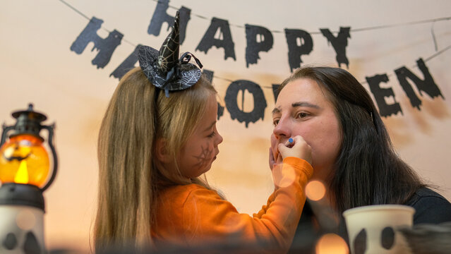 Happy little girl doing Halloween makeup while sitting at the table. Halloween celebration and festival concept. The child paints her mom