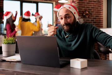 Stressed overloaded caucasian man in santa hat working on laptop at christmas season. Exhausted employee managing project deadline on computer in office during new year holiday