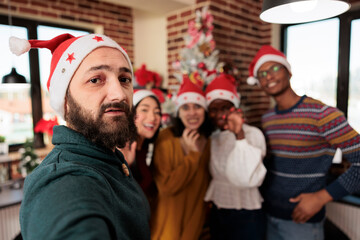 Fototapeta premium Diverse workers taking smartphone selfie at christmas corporate party in decorated office. Colleagues in santa hats posing for mobile phone photo while celebrating new year at workplace