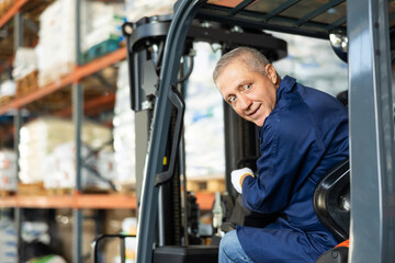 Mature man works on fork lift loader in construction rack area warehouse store repair materials. Seniore male employee looks back, chooses safe route for movement of equipment mechanism