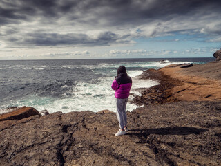 Teenager girl standing on stone surface and looking at rough stone coast with cliffs and ocean scene. Ireland, Kilkee area. Travel, tourism and sightseeing concept. Irish landscape and nature