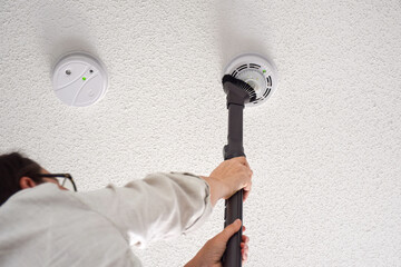 Woman vacuuming smoke and carbon monoxide detectors on a ceiling of a domestic room - obrazy, fototapety, plakaty