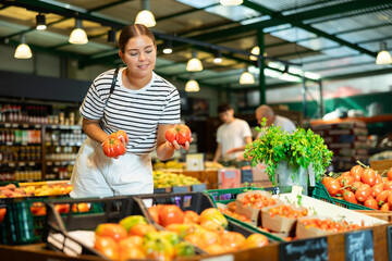 Obraz premium Interested smiling young girl shopping for organic vegetables in farm store, choosing ripe tomatoes on produce display..