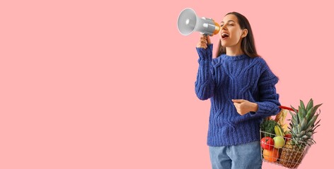 Young woman with megaphone holding shopping basket full of products on pink background with space for text