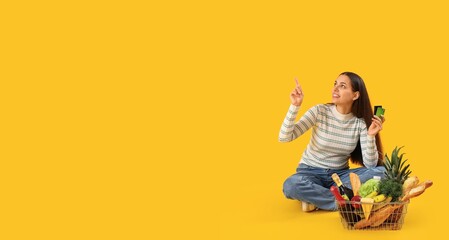 Young woman with credit cards and shopping basket full of products pointing at something on yellow background with space for text