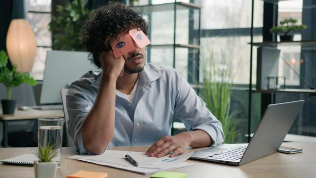 Funny lazy businessman tired male business manager napping at office desk Arabian Indian man sleep closed eyes with sticky notes comical ridiculous stickers on face sleeping guy nap pretend working