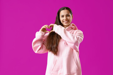 Young woman in pink hoodie making heart gesture on purple background