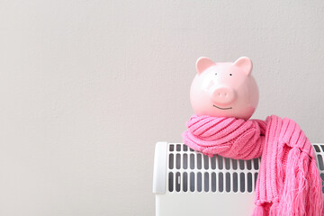 Piggy bank with warm scarf on electric convector heater near white wall at home. Heating saving concept