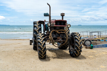 An old tractor with large wheels stands on a concrete slope into the sea. Trailers are parked nearby to pull boats out of the water and transport them.