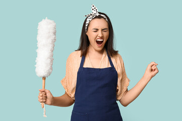 Portrait of angry young housewife in apron with pp duster on blue background - obrazy, fototapety, plakaty