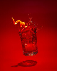 A freshly made Negroni cocktail is seen splashing dynamically against a vibrant red gradient background. A garnish of orange peel hangs above the glass.