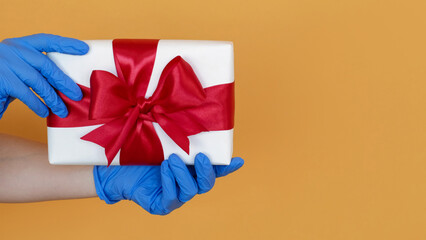 Holiday present. Christmas gift. Safety delivery. Hands in medicine blue gloves with box with red ribbon isolated on yellow background empty space.