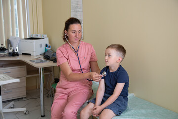 Young white woman doctor examines 5-year-old child in office of private clinic. Nurse in pink uniform with stethoscope on couch in hospital. Boy at pediatrician's appointment. Treatment, health care - obrazy, fototapety, plakaty