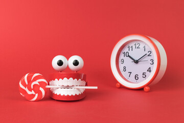 A wind-up toy with a mouth and eyes with a lollipop and a red alarm clock on a red background.