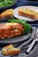 A freshly baked slice of lasagne sits on a plate beside a garden salad. Slices of garlic bread can be seen i the background.