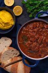 A freshly made pot of beef curry sits in a pot beside sliced bread, lemon and broccolini