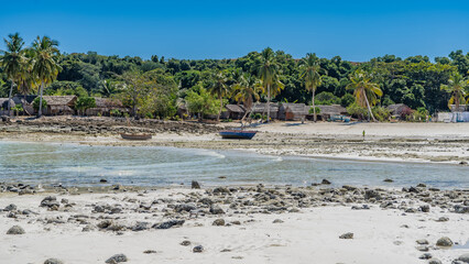 Low tide in the ocean. Stones are scattered on the exposed seabed. Wooden boats on the sand. On the shore, in the thickets of palm trees, Malagasy huts, drying laundry, tiny silhouettes of people