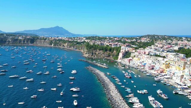 Superb aerial push-in shot from Panoramica sulla Corricella on Procida Island (Isola di Procida) of the blue Tyrrhenian Sea in the Gulf of Naples with pastel buildings, white boats and mighty Vesuvius
