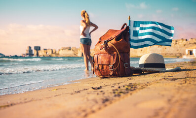 Travel destination in Greece-Traveler woman with bag, hat and Greek flag on the beach- Road trip, Adventure, summer vacation concept