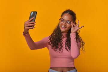 Young beautiful cheerful Indian woman student tries on glasses and takes selfie on mobile phone or uses camera of electronic gadget instead of mirror stands on orange studio background.