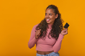 Young cheerful Indian woman zoomer shows tongue and holds credit card rejoicing at opportunity to make purchases and use cashless payments for online shopping stands posing on orange background. - obrazy, fototapety, plakaty