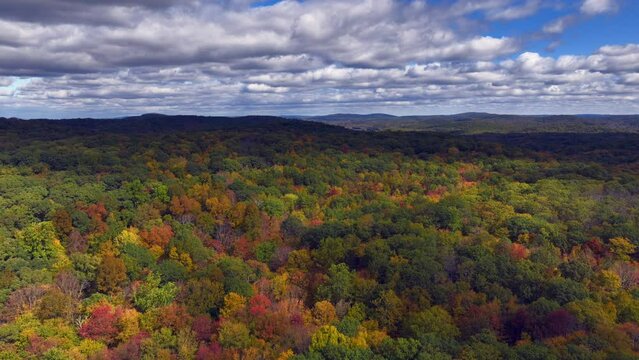 An aerial view of the mountains in Putnam County, NY on a beautiful day. The leaves of the trees are changing for the autumn season. The drone camera dolly in slowly over the colorful trees.