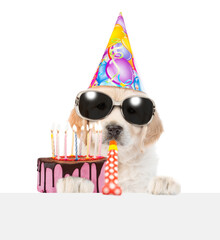 Golden retriever puppy wearing  party cap and sunglasses blows into party horn, looks above empty white banner and shows birthday cake with burning candles. isolated on white background