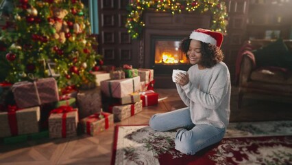 Xmas scene. Christmas, smile or happy african american woman with tea in home on holiday. Beauty young Christmas woman sitting near fireplace in dark room at home and drinking hot beverage.