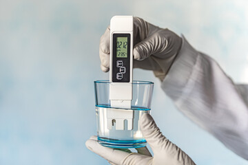 pH meter in hands with gloves, glass of water on blue background. Measurement of the characteristics of drinking water. The hardness of the water. poor water quality. high values of salt impurities - obrazy, fototapety, plakaty