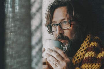 Side portrait of unhealthy man unhappy at home drinking medicine or coffee near the window looking outside. Cold interior temperature concept. Gas energy crisis problems. Flu and cold health people - obrazy, fototapety, plakaty