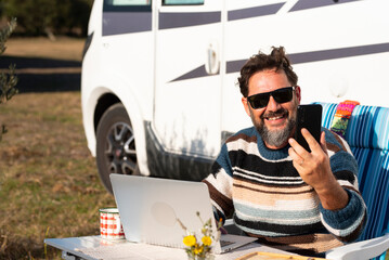 Happy traveler work on laptop computer outside a camper van and use phone call to connect and call. One man smile and enjoy cellphone calling witting outside a¡a motorhome alone at the table. Nomadic - obrazy, fototapety, plakaty