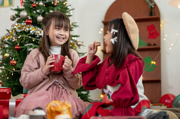 Two joyful Asian girls are enjoying hot chocolate and cookies and celebrating Christmas at home.
