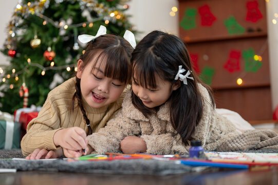 A cute Asian girl is helping her sister making a Christmas card while lying on the floor together.