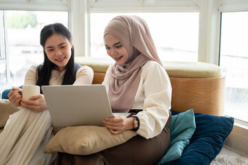 An Asian-Muslim woman is watching videos or a movie on a laptop with her friend in a room together.