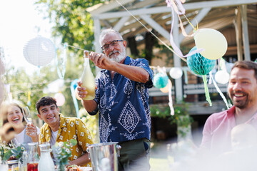 Grandfather opening bottle of champagne, pouring a champagne into glasses. Senior man making celebratory toast at outdoor summer garden party. - obrazy, fototapety, plakaty
