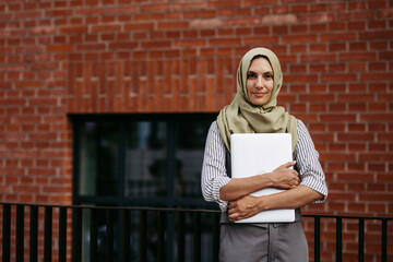 Beautiful woman in hijab standing on city street. Muslim businesswoman with laptop in hands. Iran, Afganistan female teacher in front of school building.