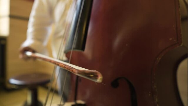 extreme close up of weary used scratched cello bass being played slowly by a professional musician inside building with wooden walls little round seat next to him man wearing white shirt grey pants
