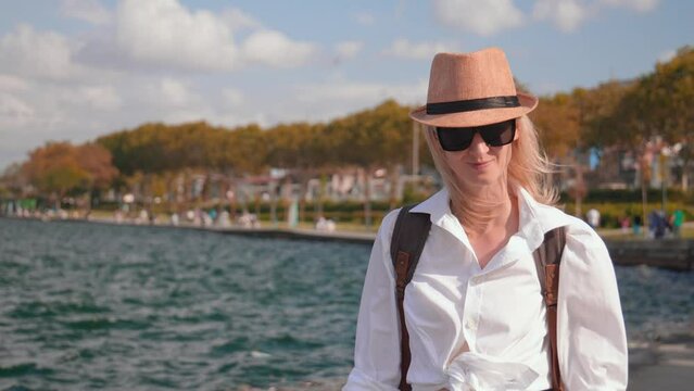 Young beautiful woman with backpack in sunglasses walks along promenade in Istanbul in Turkey and wind blows her hat off, she adjusts it with her hand. Portrait of tourist girl with smile.