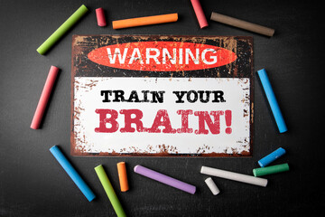 Train Your Brain. Metal warning sign and colored pieces of chalk on a dark chalkboard background