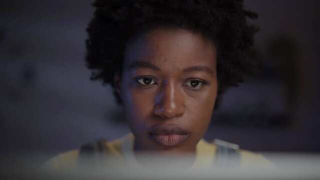 Close-up of concentrated young adult afro woman staring at computer screen indoor at night. African American female in darkened room using laptop. Millennial people working from home after hours. 