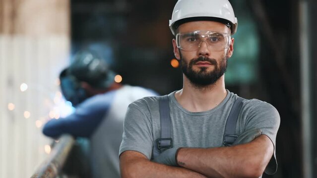 Portrait of a professional heavy industry engineer. Worker wearing protective uniform, goggles and helmet, smiling. In the background is a disoriented large industrial plant where welding sparks fly.