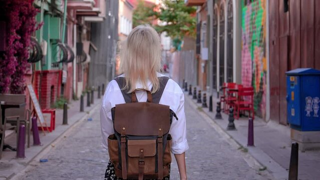 Slow motion beautiful female tourist with backpack in white shirt and straw hat walking along empty street in old tourist part city, enjoying her vacation. Sightseeing in popular tourist destination.