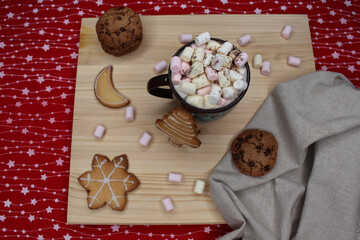 Brown cup or mug of coffee or hot chocolate with marshmallows and cocoa powder. Cookies with chocolate chips and with Christmas shapes. A gray kitchen rag. A light wood and a red and white tablecloth.
