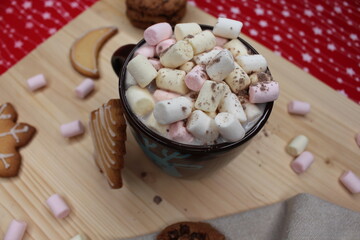 Brown cup or mug of coffee or hot chocolate with marshmallows and cocoa powder. Cookies with chocolate chips and with Christmas shapes. A gray kitchen rag. A light wood and a red and white tablecloth.