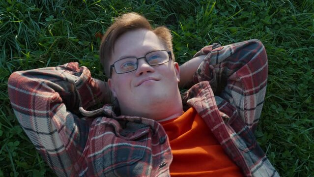 Portrait of young man with down syndrome in checkered shirt laying on the grass outdoors in the public park. Man with trisomy 21 illness. Disabled guy having a rest