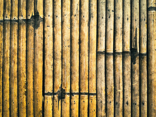 A close-up photo of a yellow and brown bamboo fence, arranged in vertical lines, with a weathered and rustic appearance